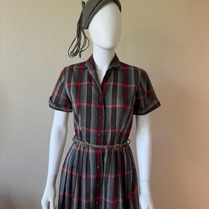 Peut inclure: Une robe vintage &agrave; carreaux gris, rouges et noirs. La robe a des manches courtes, une fermeture boutonn&eacute;e sur le devant et une ceinture assortie. Un b&eacute;ret gris avec une pampille est &eacute;galement inclus. La robe est pr&eacute;sent&eacute;e sur un mannequin.