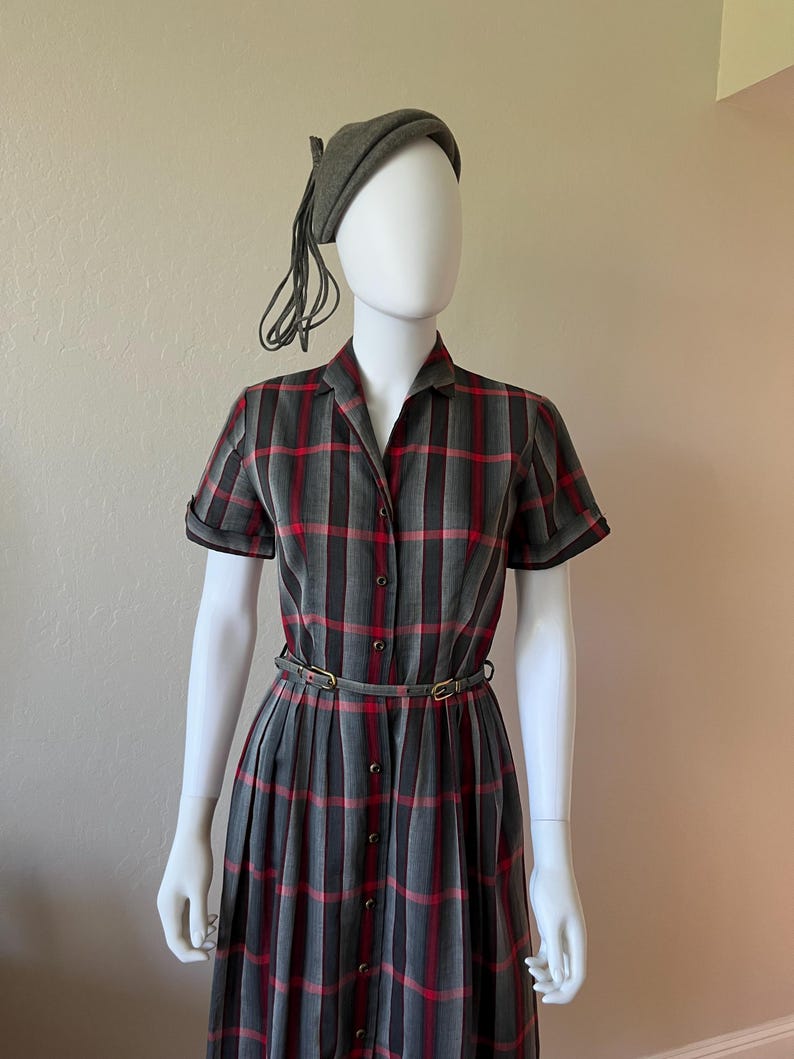Peut inclure: Une robe vintage &agrave; carreaux gris, rouges et noirs. La robe a des manches courtes, une fermeture boutonn&eacute;e sur le devant et une ceinture assortie. Un b&eacute;ret gris avec une pampille est &eacute;galement inclus. La robe est pr&eacute;sent&eacute;e sur un mannequin.