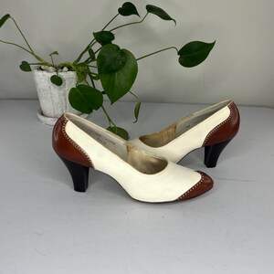 May include: A pair of vintage women's heels. The shoes are white with brown accents on the toe, heel, and trim. The heels are dark brown and the shoes have a classic, retro design. The heels are approximately 2.5 inches tall.