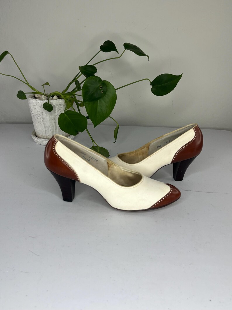 May include: A pair of vintage women's heels. The shoes are white with brown accents on the toe, heel, and trim. The heels are dark brown and the shoes have a classic, retro design. The heels are approximately 2.5 inches tall.