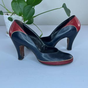 Könnte beinhalten: Ein Paar Vintage-Pumps mit hohen Absätzen. Die Schuhe sind marineblau mit roten Akzenten an Ferse und Zehen sowie einem goldenen Besatz. Die Absätze sind dunkelbraun. Die Schuhe werden auf einer hellblauen Oberfläche präsentiert.