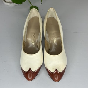 May include: A pair of vintage cream-colored high heels with brown leather toe caps. The shoes have a classic design with a pointed toe and a stiletto heel. The interior of the shoes has text that reads "Hills and Dale" and "Steinbach Co".
