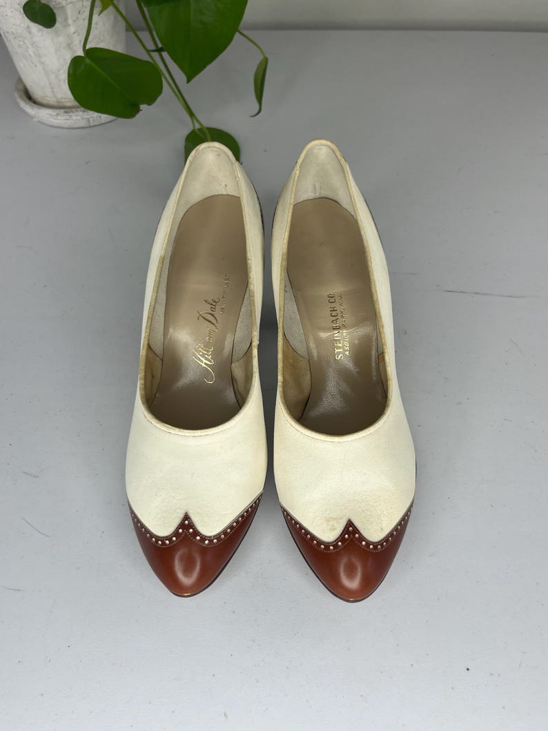 May include: A pair of vintage cream-colored high heels with brown leather toe caps. The shoes have a classic design with a pointed toe and a stiletto heel. The interior of the shoes has text that reads "Hills and Dale" and "Steinbach Co".