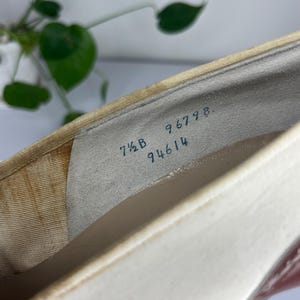 May include: Close-up of a white and beige shoe interior. The shoe has a cream-colored exterior and a dark heel. The size is 7 1/2 B, with the numbers 96798 and 94614 visible inside the shoe.