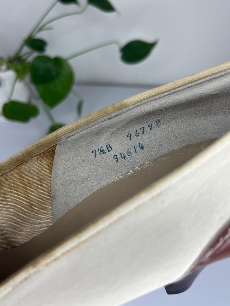May include: Close-up of a white and beige shoe interior. The shoe has a cream-colored exterior and a dark heel. The size is 7 1/2 B, with the numbers 96798 and 94614 visible inside the shoe.