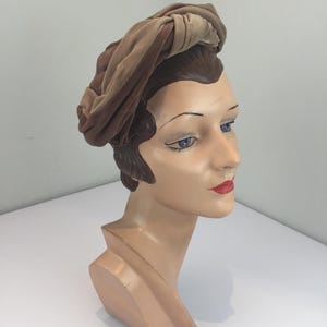 May include: A tan and brown velvet turban-style hat featuring a knotted design on the top. The hat is displayed on a stylized mannequin head with brown hair, blue eyes, and red lips, showcasing the soft texture and layered fabric construction.