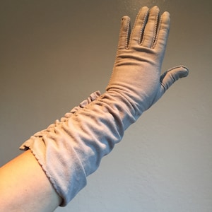 Könnte beinhalten: Ein Paar lange, hellgraue Handschuhe mit einem gerafften, gebauschten Bündchen. Die Handschuhe sind aus einem weichen, dehnbaren Stoff.