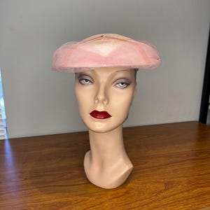 May include: A light pink hat featuring a structured crown and a wide, flat brim. The hat is adorned with a delicate, fine-mesh netting that overlays the crown and extends slightly beyond the brim, creating a soft, layered texture across the surface.