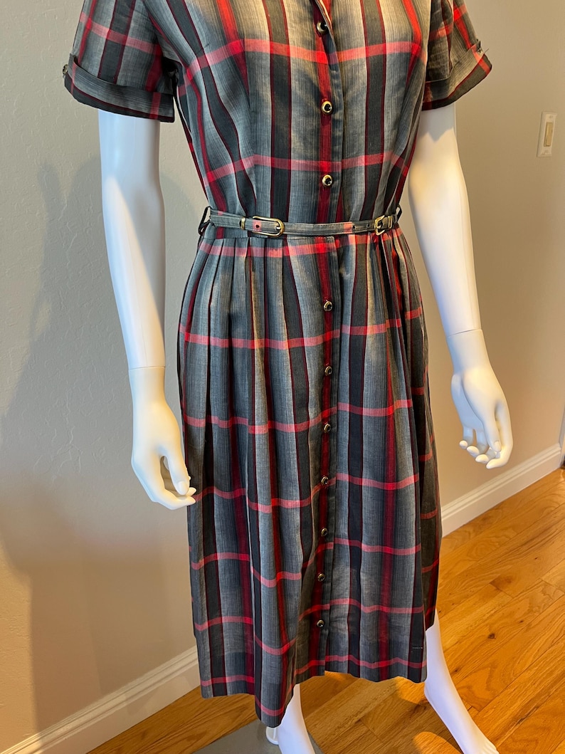 Peut inclure: Robe &agrave; carreaux grise, rouge et noire, &agrave; manches courtes et &agrave; boutons sur le devant. La robe est cintr&eacute;e &agrave; la taille par une fine ceinture. La robe est pr&eacute;sent&eacute;e sur un mannequin.