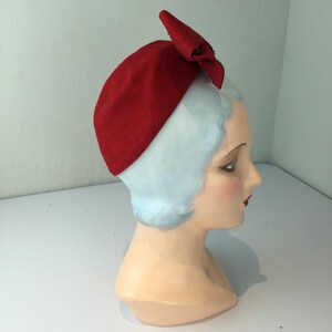 May include: A red felt hat with a bow detail. The hat is displayed on a mannequin head with light blue hair and a fair complexion. The hat is a solid, vibrant red color.