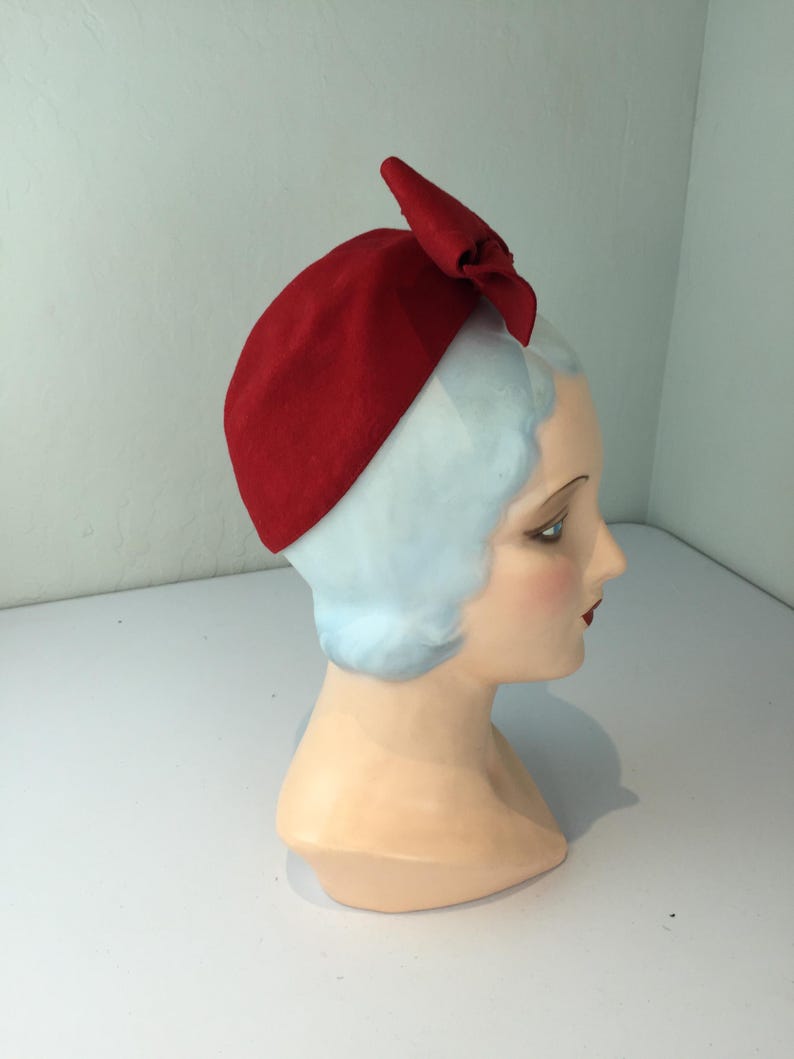 May include: A red felt hat with a bow detail. The hat is displayed on a mannequin head with light blue hair and a fair complexion. The hat is a solid, vibrant red color.