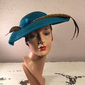May include: A teal blue felt hat with a brown pheasant feather accent. The hat has a wide brim and a slightly tilted crown.