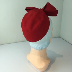 May include: A red felt hat with a large bow on top. The hat is a deep shade of red and is displayed on a mannequin head. The bow is a prominent feature, adding a touch of elegance to the hat's design. The hat is a vintage style.