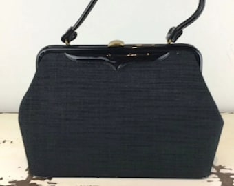 Puede incluir: Un bolso de mano de tela negra con ribetes de charol negro y un cierre dorado. El bolso de mano tiene un asa superior.