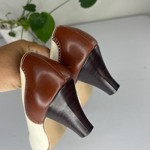 May include: A pair of two-tone leather heels. The shoes feature a brown leather heel and upper, with a cream-colored toe and side panel. The heels are dark brown and have a unique, angled shape. The shoes are held up by a hand.