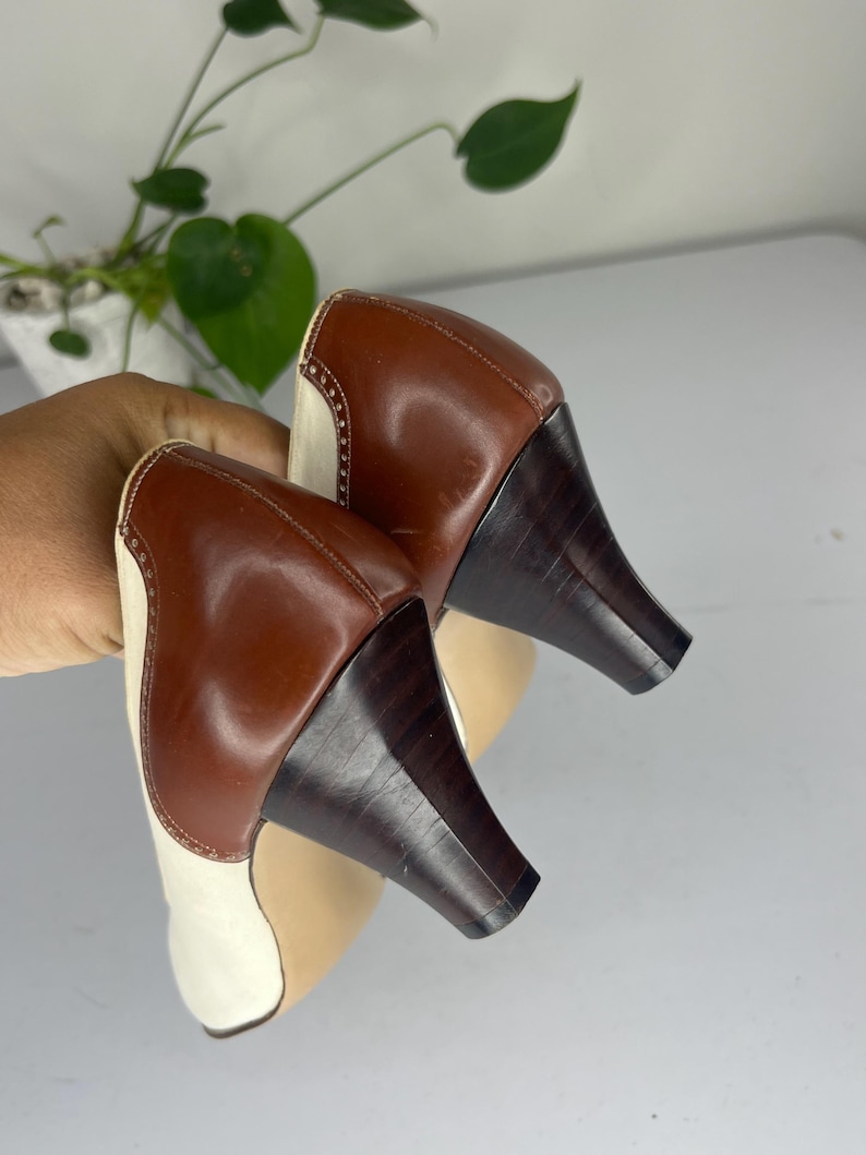 May include: A pair of two-tone leather heels. The shoes feature a brown leather heel and upper, with a cream-colored toe and side panel. The heels are dark brown and have a unique, angled shape. The shoes are held up by a hand.