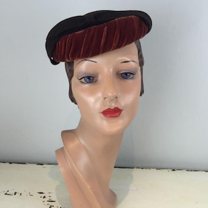 Puede incluir: Un sombrero vintage con una corona negra y un ala de terciopelo plisado de color óxido. El sombrero se exhibe en una cabeza de maniquí con ojos azules y lápiz labial rojo, mostrando un estilo retro clásico.