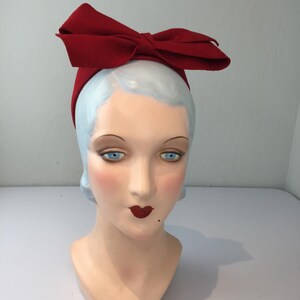 May include: A red bow headband is displayed on a mannequin head with blue eyes, red lips, and a beauty mark. The headband is a vibrant red color and features a large bow on top.