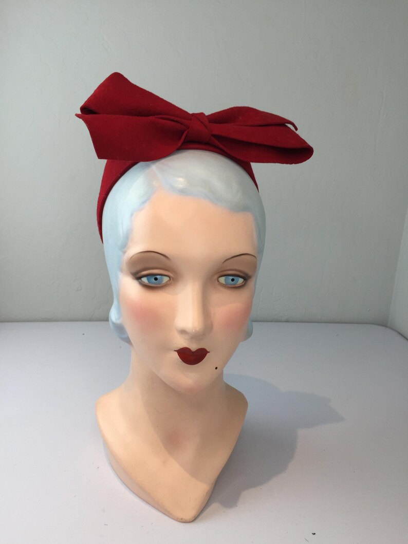 May include: A red bow headband is displayed on a mannequin head with blue eyes, red lips, and a beauty mark. The headband is a vibrant red color and features a large bow on top.