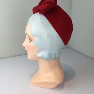May include: A vintage red hat with a large bow on top, displayed on a mannequin head. The hat is a solid, vibrant red color and appears to be made of felt or a similar material. The mannequin head has pale skin, blue hair, and red lips.