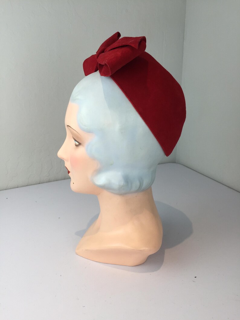 May include: A vintage red hat with a large bow on top, displayed on a mannequin head. The hat is a solid, vibrant red color and appears to be made of felt or a similar material. The mannequin head has pale skin, blue hair, and red lips.