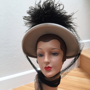 May include: A vintage-style hat with a wide brim, adorned with a large cluster of black feathers. The hat is a light tan color and features a black netting veil. The hat is displayed on a mannequin head.
