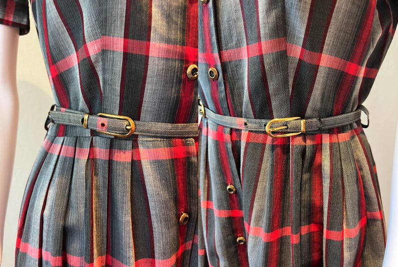 Peut inclure: Gros plan de deux robes &agrave; motif &agrave; carreaux gris, rouges et noirs. Chaque robe a une ceinture assortie avec une boucle dor&eacute;e. Les robes ont un devant boutonn&eacute; et des manches courtes. Les robes sont probablement de style vintage ou r&eacute;tro.