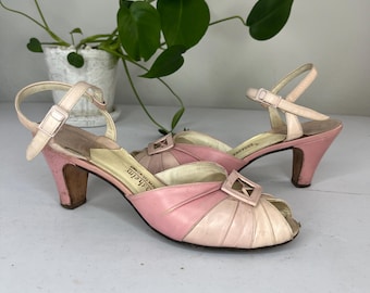 May include: A pair of pink and white heeled sandals with a buckle detail. The sandals are made of leather and have a peep toe design. The brand name "Saladin" is printed on the insole.