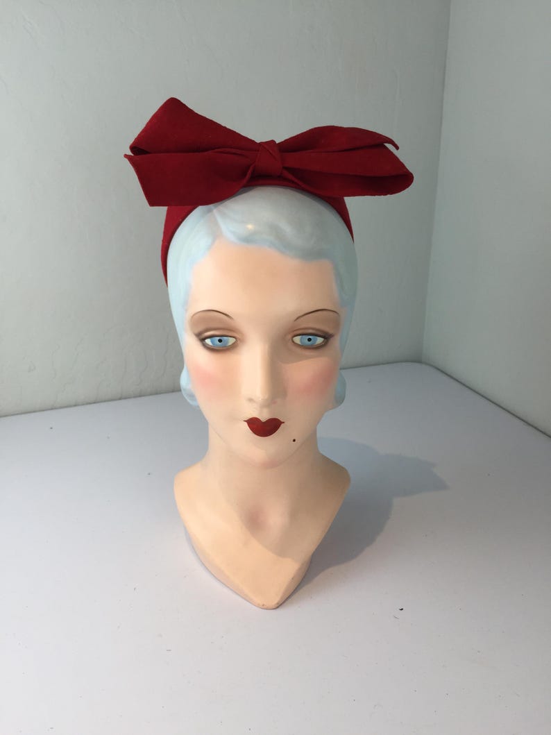May include: A red bow headband on a mannequin head. The headband features a large, structured bow. The mannequin has pale skin, blue eyes, red lips, and light blue hair.