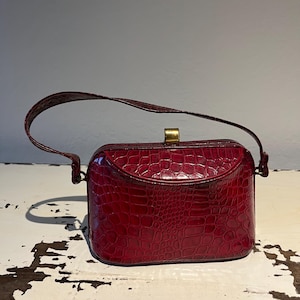 Peut inclure: Un sac à main rouge foncé avec un motif texturé gaufré façon crocodile, doté d'un corps rectangulaire structuré, d'une anse unique arquée et d'un fermoir en métal doré centré sur le bord supérieur de la structure du sac.