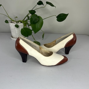 May include: A pair of vintage women's heels. The shoes are white with brown accents on the toe and heel. The heels are dark brown and the shoes have a classic, retro design.