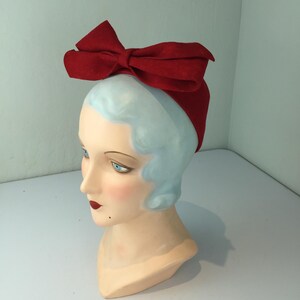 May include: A red bow headband is displayed on a mannequin head with blue hair and a fair complexion. The bow is large and prominent, made of a textured material. The mannequin has blue eyes and red lipstick.