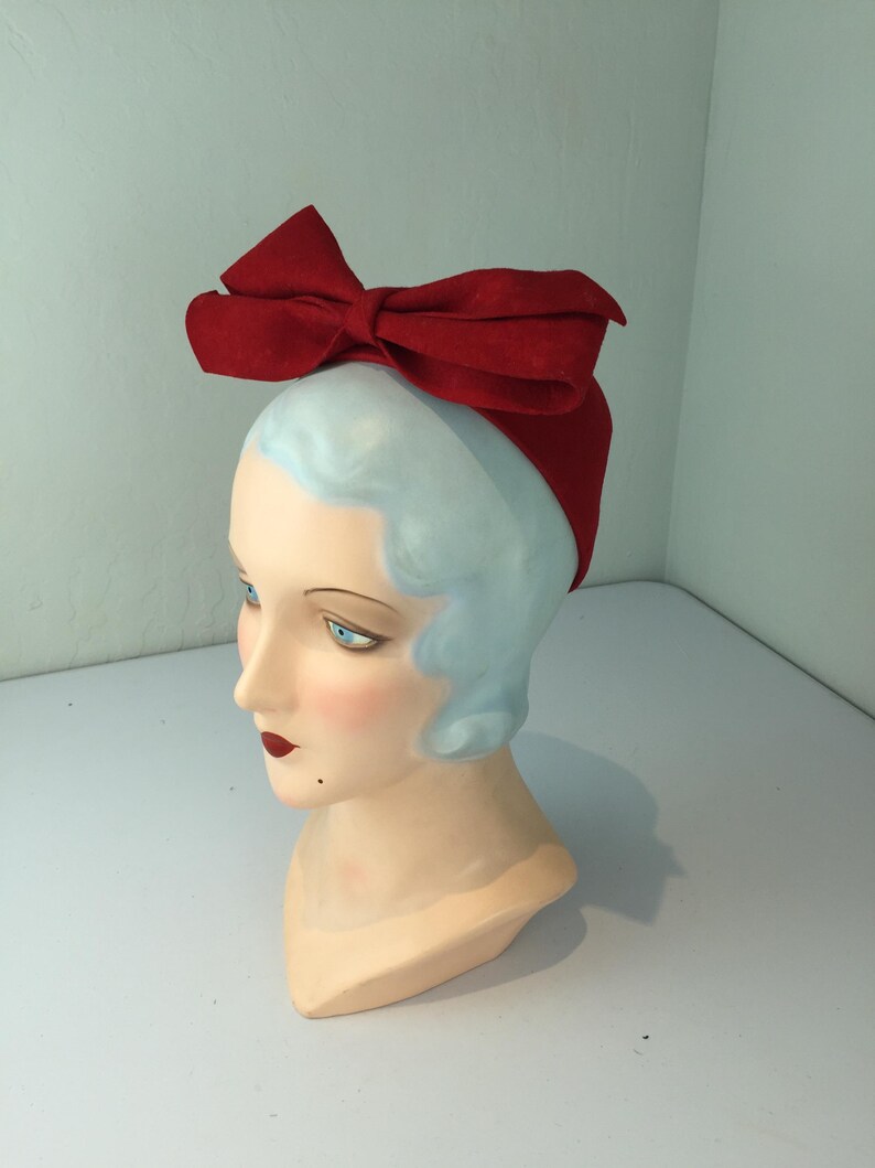 May include: A red bow headband is displayed on a mannequin head with blue hair and a fair complexion. The bow is large and prominent, made of a textured material. The mannequin has blue eyes and red lipstick.