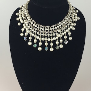 May include: A silver chain necklace with multiple strands of faux pearls and iridescent crystal accents. The necklace has a cascading design with the pearls hanging in a tiered fashion.