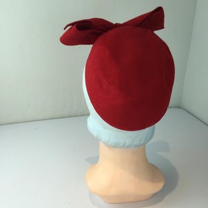 May include: A red felt hat with a bow detail. The hat is displayed on a mannequin head with a light blue neck and a peach-colored face. The hat's design is simple and classic.