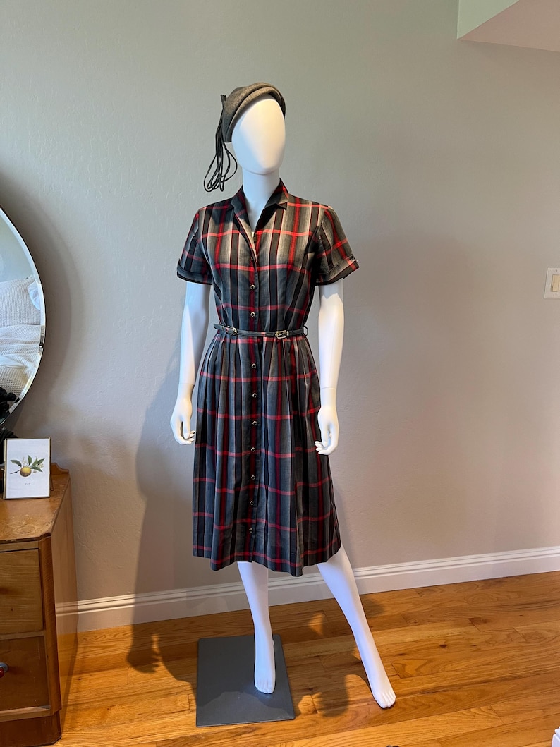 Peut inclure: Une robe vintage &agrave; carreaux gris, rouges et noirs, pr&eacute;sent&eacute;e sur un mannequin. La robe a des manches courtes, un devant boutonn&eacute; et une ceinture assortie. Un b&eacute;ret gris compl&egrave;te la tenue. La robe arrive au genou.