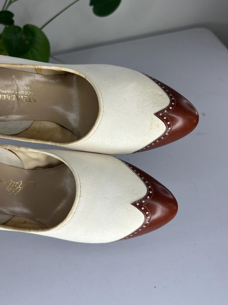 May include: Pair of vintage women's shoes. The shoes are two-tone, with white uppers and brown leather toe caps. The toe caps have decorative perforations. The shoes are in good condition, with some signs of wear. The brand name "Steinbach" is visible inside the shoe.