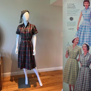 Peut inclure: Une robe &agrave; carreaux de style vintage en gris, rouge et noir, pr&eacute;sent&eacute;e sur un mannequin. La robe pr&eacute;sente des manches courtes, une fermeture boutonn&eacute;e sur le devant et une ceinture assortie. Un chapeau assorti est &eacute;galement sur le mannequin. Une publicit&eacute; vintage pour des robes est en arri&egrave;re-plan.