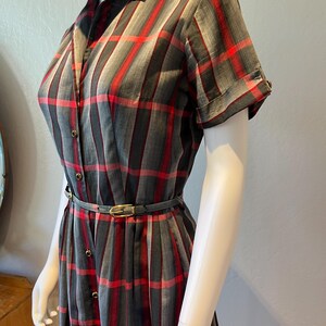 Peut inclure: Robe vintage &agrave; carreaux gris, rouges et noirs, boutonn&eacute;e sur le devant. Elle pr&eacute;sente des manches courtes, un col chemise et une ceinture assortie avec une boucle dor&eacute;e. La robe est pr&eacute;sent&eacute;e sur un mannequin.