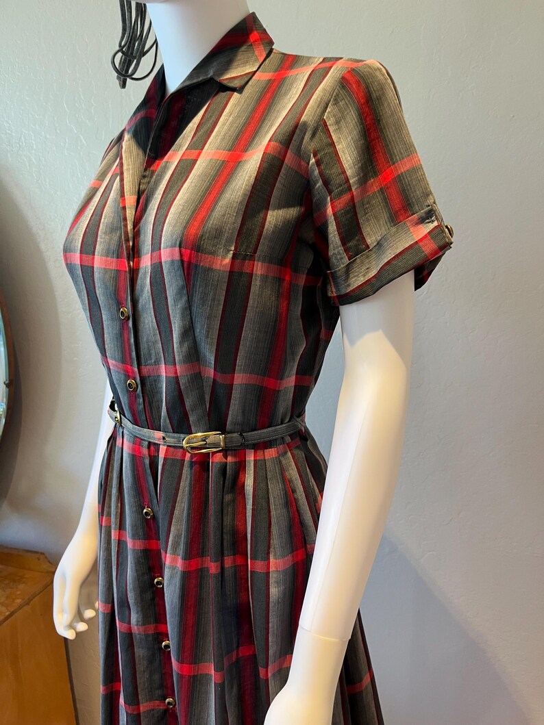 Peut inclure: Robe vintage &agrave; carreaux gris, rouges et noirs, boutonn&eacute;e sur le devant. Elle pr&eacute;sente des manches courtes, un col chemise et une ceinture assortie avec une boucle dor&eacute;e. La robe est pr&eacute;sent&eacute;e sur un mannequin.