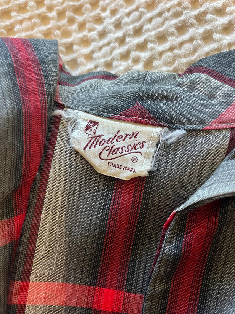 Peut inclure: Gros plan sur une chemise vintage &agrave; rayures grises, rouges et noires. La chemise pr&eacute;sente un col et une &eacute;tiquette indiquant "Modern Classics TRADE MARK". Le tissu a un aspect doux et textur&eacute;.
