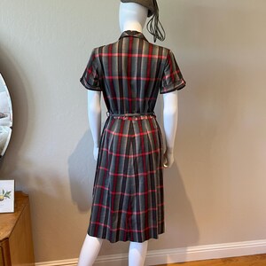 Peut inclure: Une robe vintage &agrave; carreaux gris, rouges et noirs, pr&eacute;sent&eacute;e sur un mannequin. La robe pr&eacute;sente des manches courtes, une taille ceintur&eacute;e et une jupe pliss&eacute;e. Un b&eacute;ret assorti avec un pompon est &eacute;galement inclus. La robe est de style classique.
