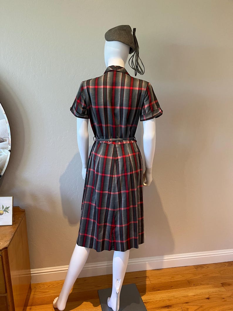 Peut inclure: Une robe vintage &agrave; carreaux gris, rouges et noirs, pr&eacute;sent&eacute;e sur un mannequin. La robe pr&eacute;sente des manches courtes, une taille ceintur&eacute;e et une jupe pliss&eacute;e. Un b&eacute;ret assorti avec un pompon est &eacute;galement inclus. La robe est de style classique.