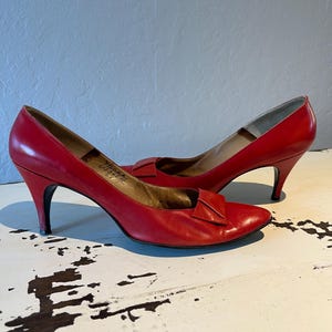May include: A pair of red leather pumps with pointed toes and small heels. Each shoe has a decorative bow on the vamp. The heels are black. The shoes are on a distressed white surface.