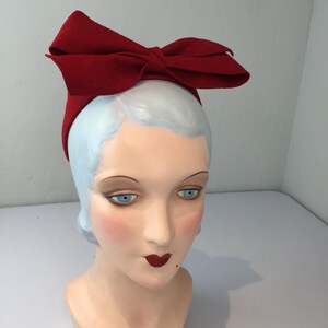 May include: A red bow headband is displayed on a mannequin head. The headband features a large, structured bow made of a solid red fabric. The mannequin head has blue eyes, red lips, and a pale complexion, creating a vintage aesthetic.