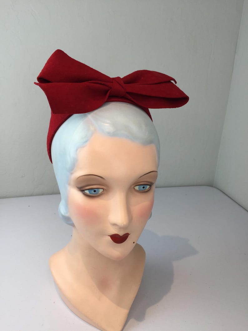 May include: A red bow headband is displayed on a mannequin head. The headband features a large, structured bow made of a solid red fabric. The mannequin head has blue eyes, red lips, and a pale complexion, creating a vintage aesthetic.