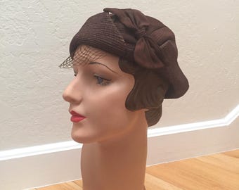 Puede incluir: Un sombrero cloche marrón vintage con un lazo y un delicado velo de red. El sombrero se exhibe en una cabeza de maniquí, mostrando su forma y diseño. El color del sombrero es un marrón oscuro intenso y parece estar hecho de un material tejido.
