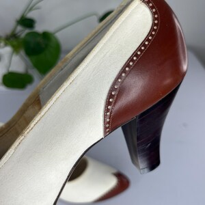 May include: Vintage two-tone heels in white and brown leather. The shoes feature a classic design with a rounded toe, a sturdy heel, and decorative stitching along the edges. The heels are approximately 3 inches high.