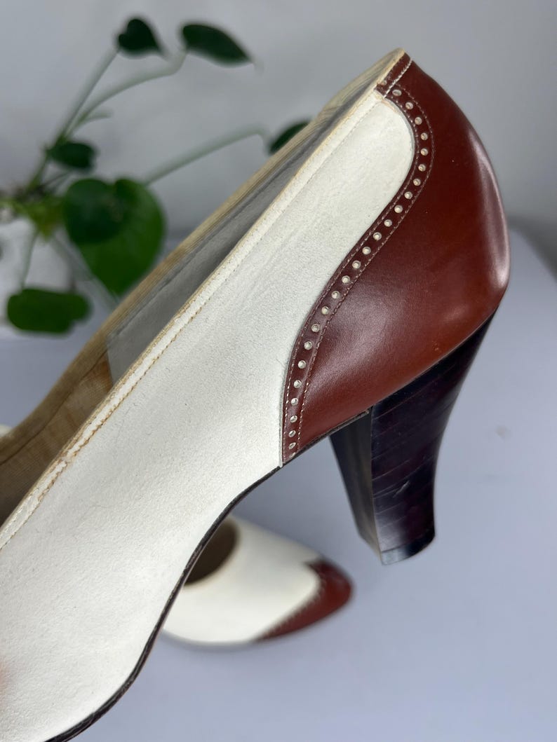 May include: Vintage two-tone heels in white and brown leather. The shoes feature a classic design with a rounded toe, a sturdy heel, and decorative stitching along the edges. The heels are approximately 3 inches high.
