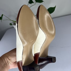 May include: A pair of vintage women's heels, viewed from the soles. The shoes feature a white upper with brown accents on the toe and heel. The heels are a dark brown, and the soles are a light tan color. The shoes have a classic, elegant design.